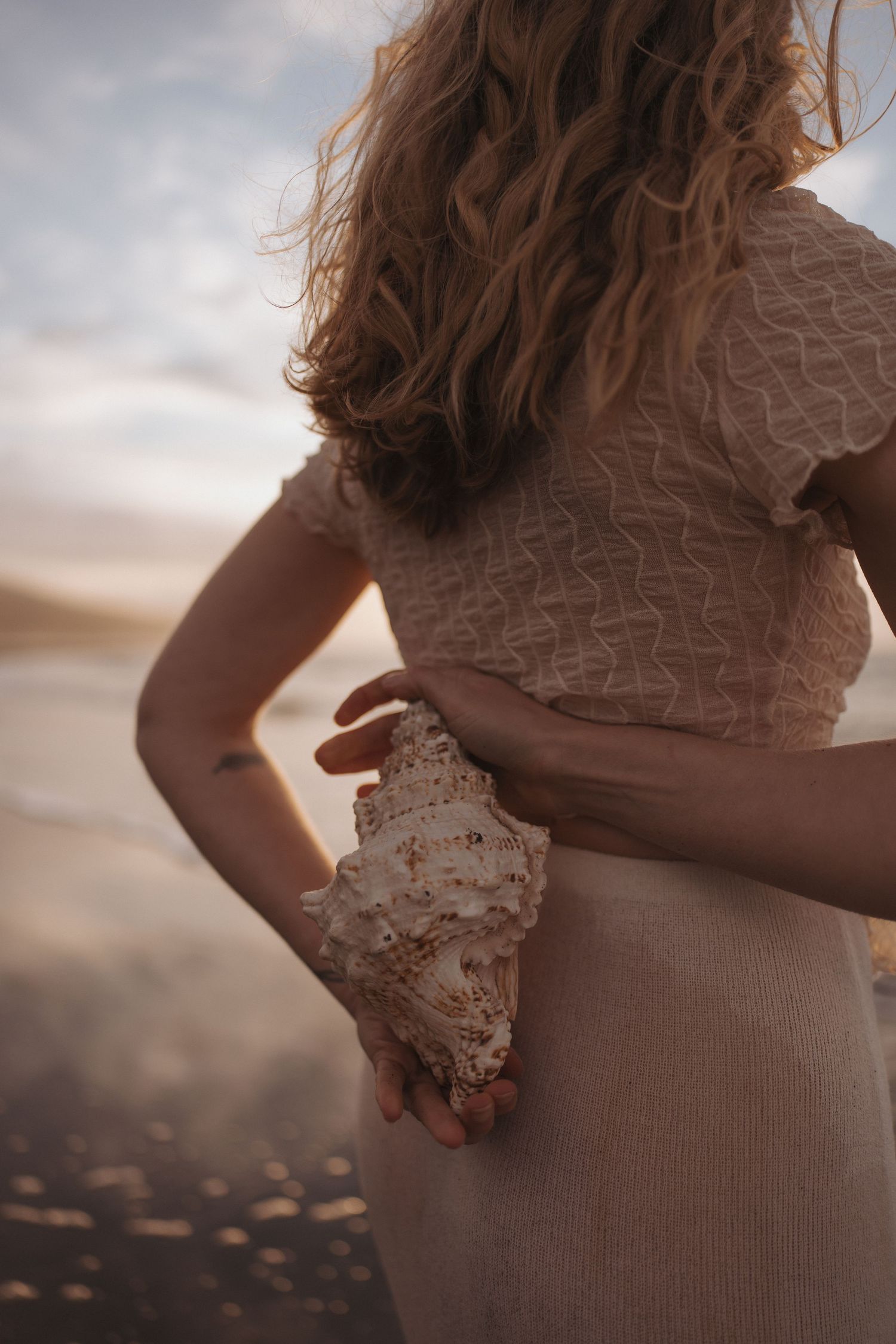 Woman with wavy hair in a knit cream dress holds a large spiral seashell behind her back on a beach at sunset.