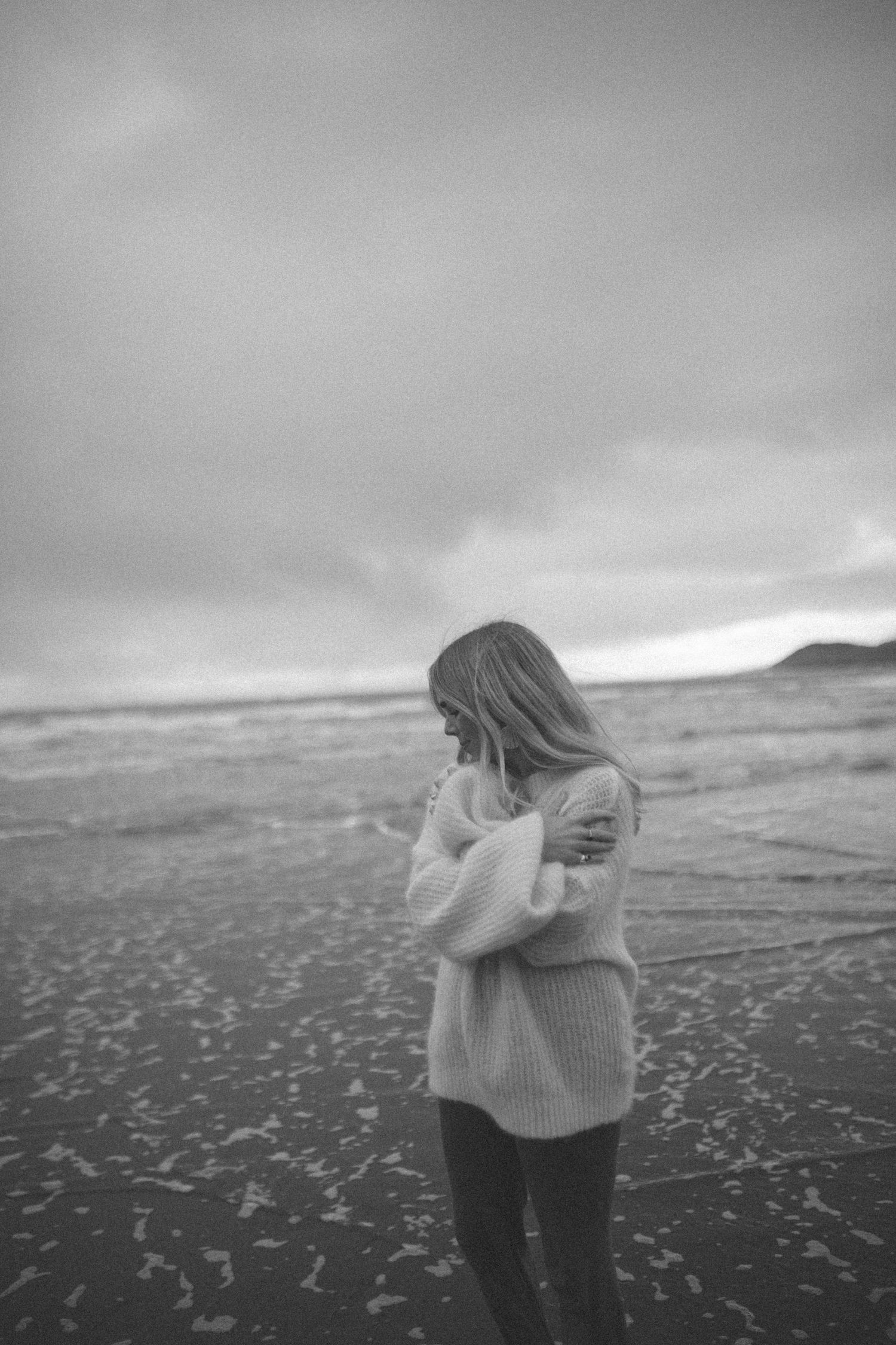 Woman in a chunky knit sweater hugging herself on a windswept beach under a moody overcast sky, in black and white.
