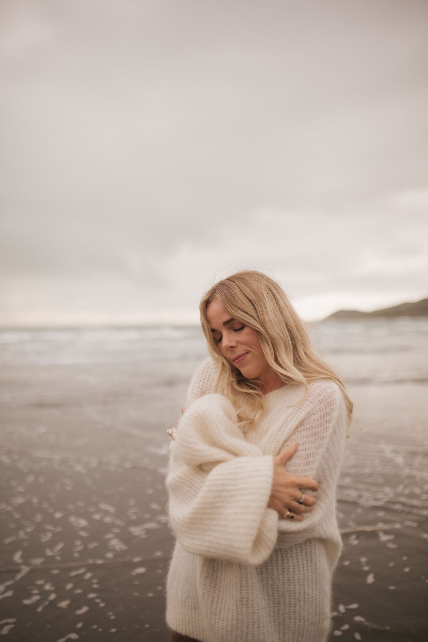 Woman with long blonde hair hugging herself in a cozy cream knit sweater on a moody, overcast beach with gentle waves.