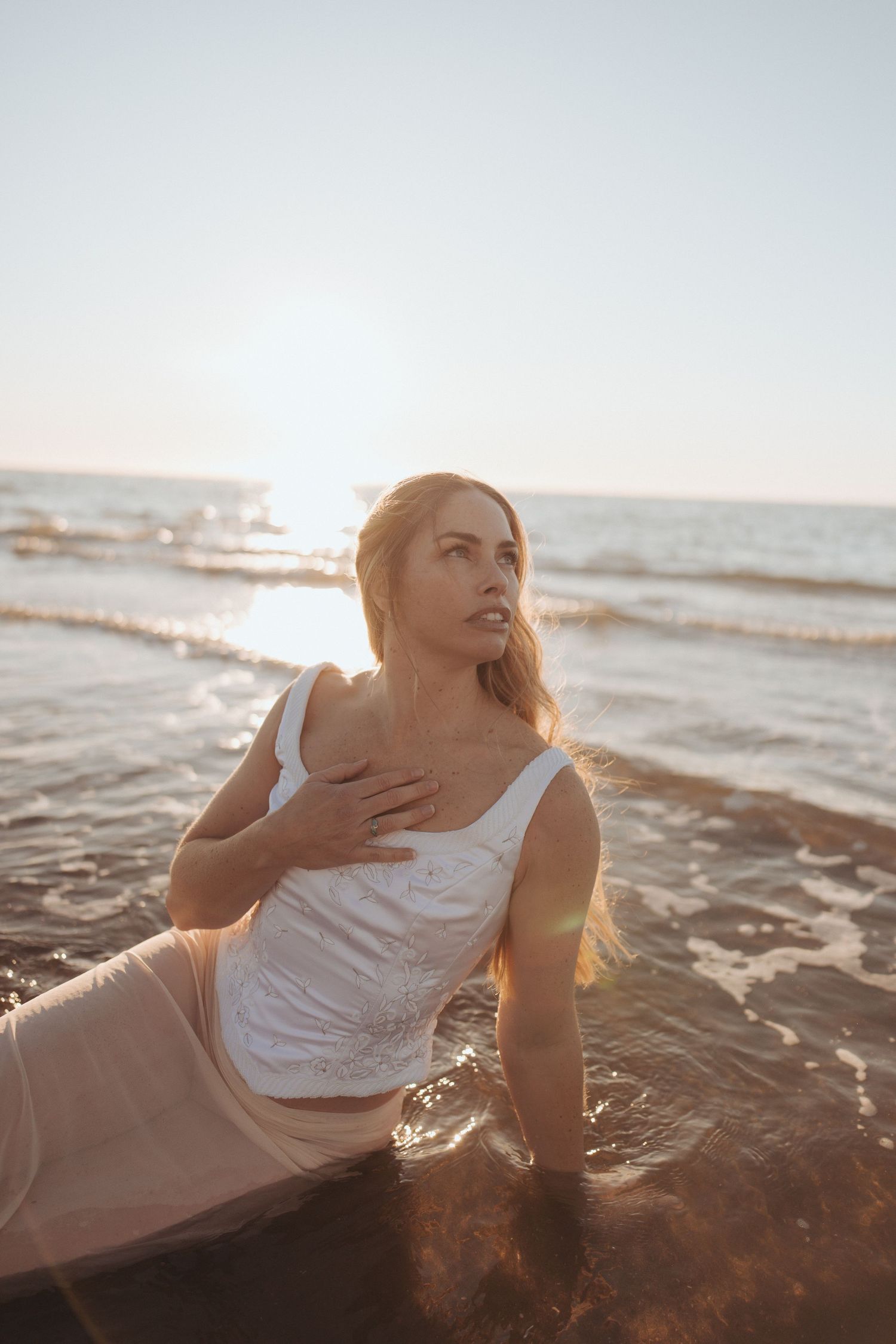 Woman with long blonde hair in a white lace top sits in shallow ocean waves at golden hour, gazing upward with her hand on her chest.