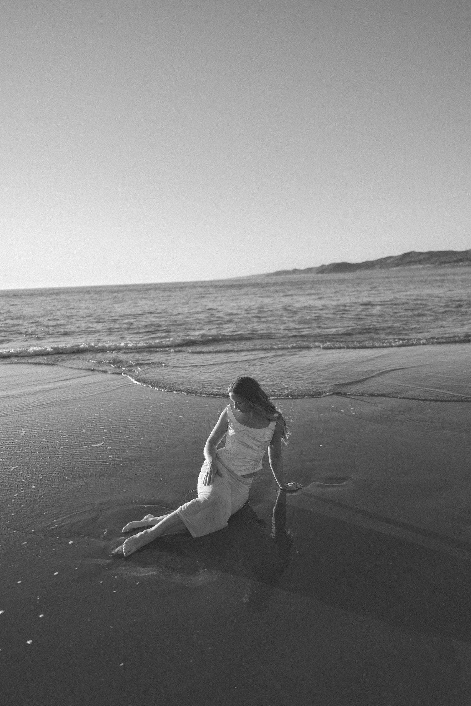 Black and white photo of a woman in a light dress sitting on a wet sandy beach at the water's edge with gentle waves and distant hills.