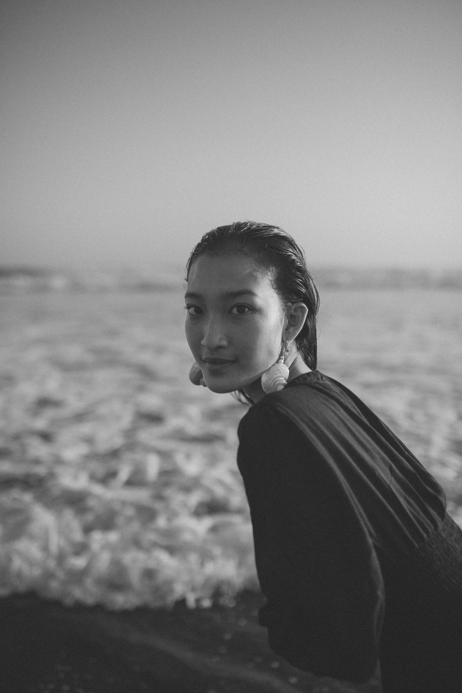 Black and white portrait of a woman with wet slicked-back hair and pearl earrings glancing over her shoulder by crashing ocean waves.