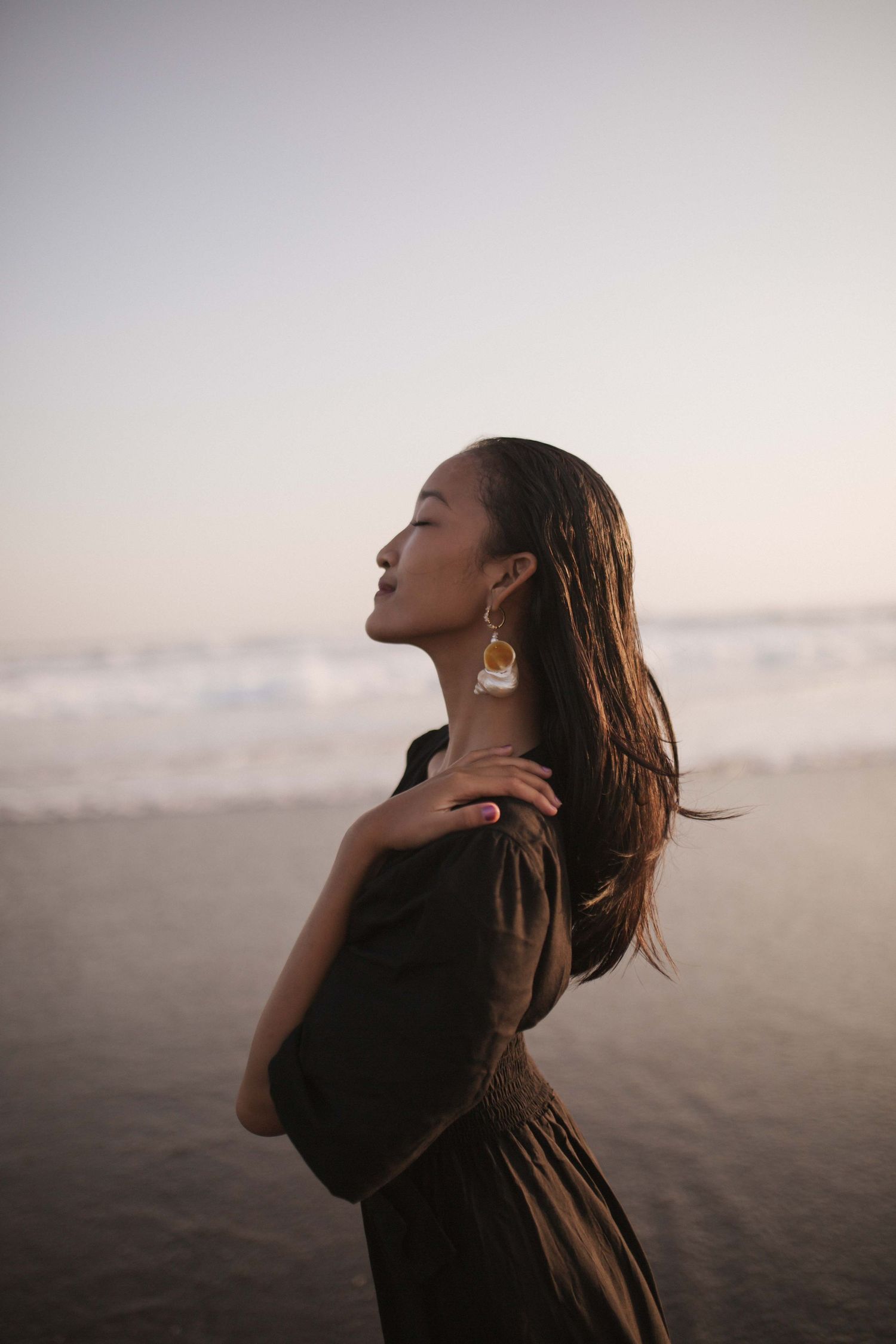 Woman with wet dark hair and gold drop earrings stands on a beach at sunset, eyes closed, wearing a black dress with waves behind her.