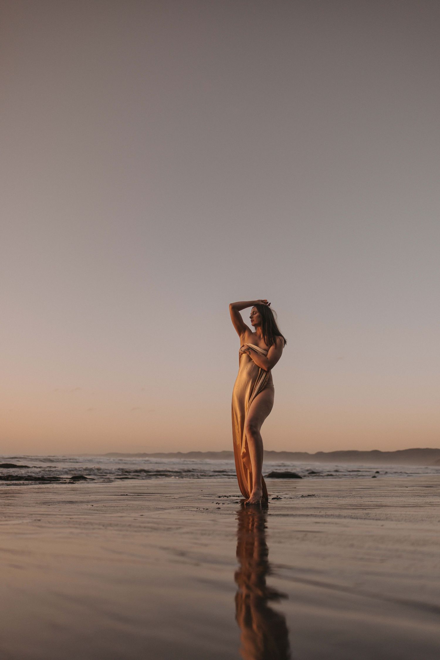 Woman in a flowing golden dress poses on a wet sandy beach at sunset, her reflection shimmering in the shallow water under a soft peach sky.