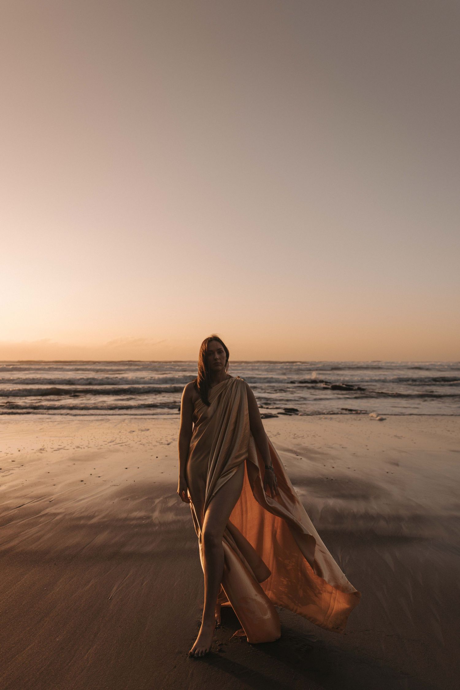 Woman in a flowing golden satin wrap stands barefoot on a wet sandy beach at sunset, with gentle waves and a warm glowing sky behind her.