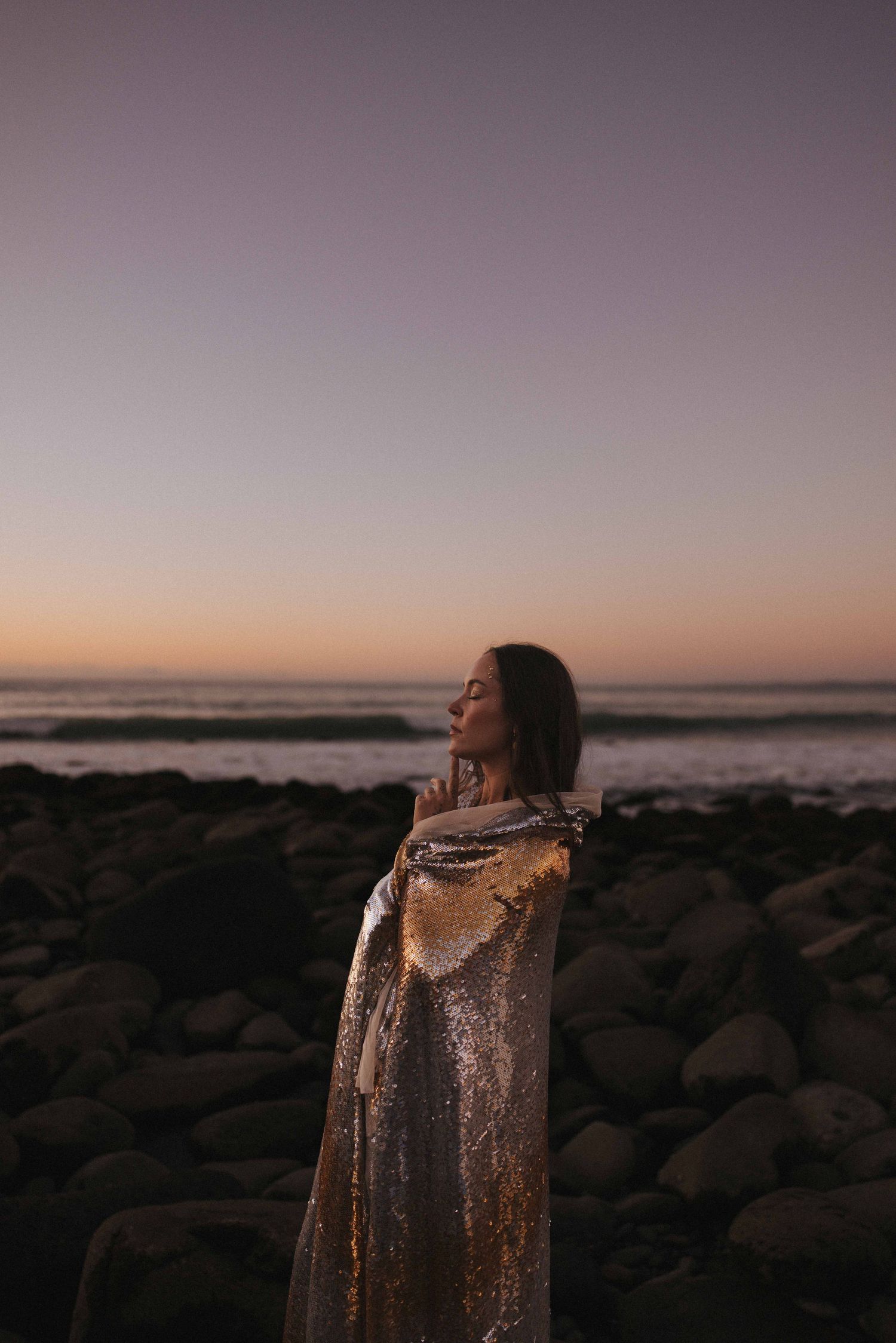 Woman wrapped in a shimmering sequin cloth stands with closed eyes on a rocky beach at dusk, with a pastel sunset sky over the ocean.