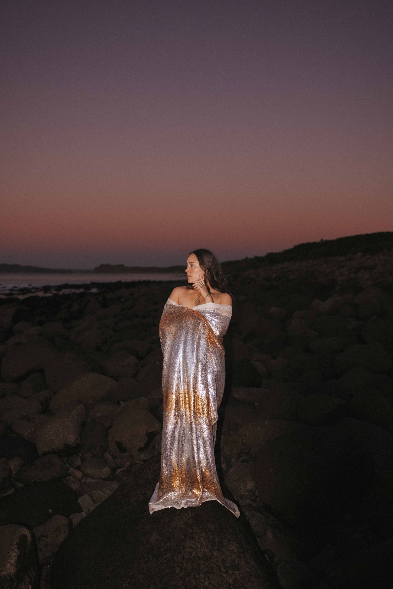 Woman wrapped in a shimmering metallic gold fabric stands on rocky shoreline at dusk beneath a purple and pink twilight sky.