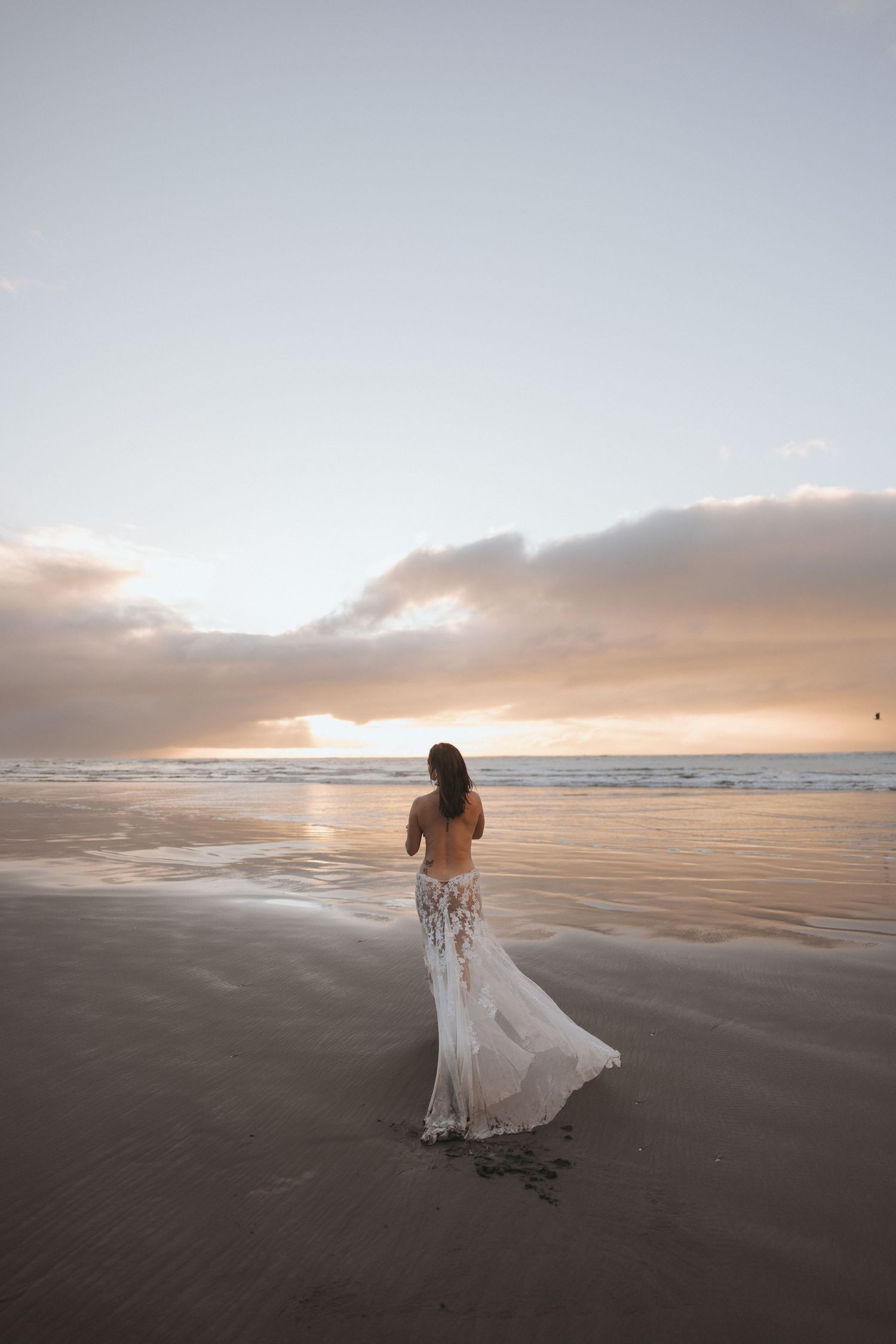 Woman in a flowing white lace skirt stands on a wet sandy beach at sunset, gazing at the glowing horizon beneath soft clouds.