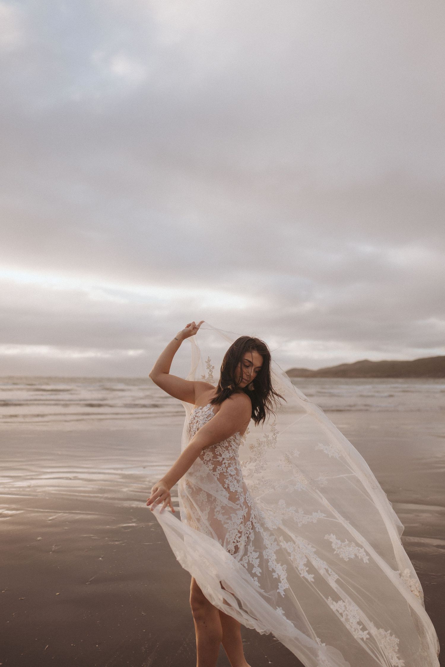 Woman in a sheer floral lace gown twirls on a wet sandy beach at sunset, her long lace veil flowing in the wind.