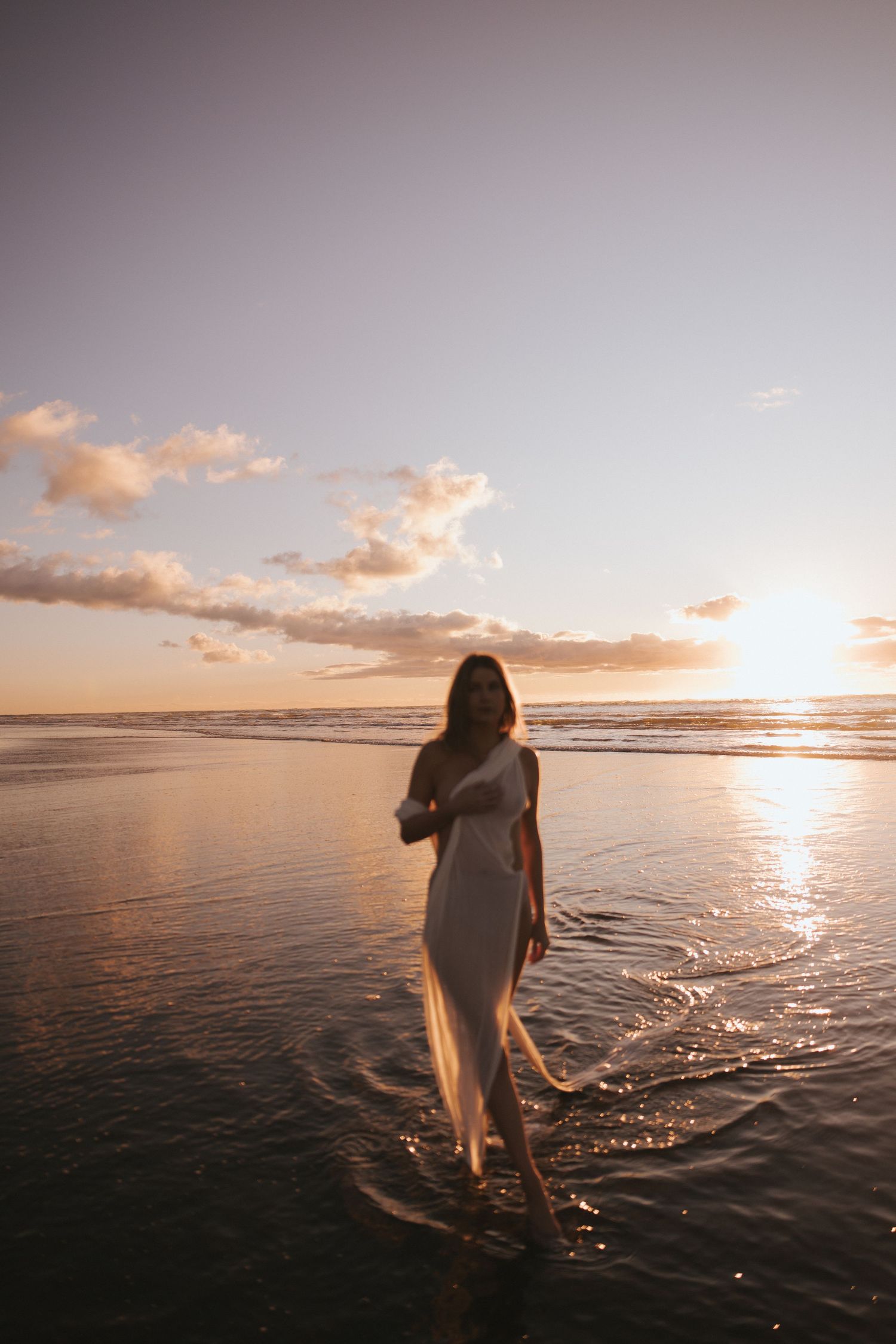 Woman in a flowing white dress stands in shallow ocean water at sunset, with golden light reflecting on the wet sand and waves.