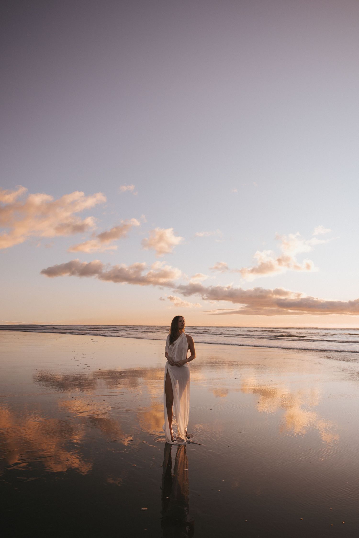 Woman in a flowing white dress stands on a wet beach at sunset, with pastel clouds reflected in the glassy sand.