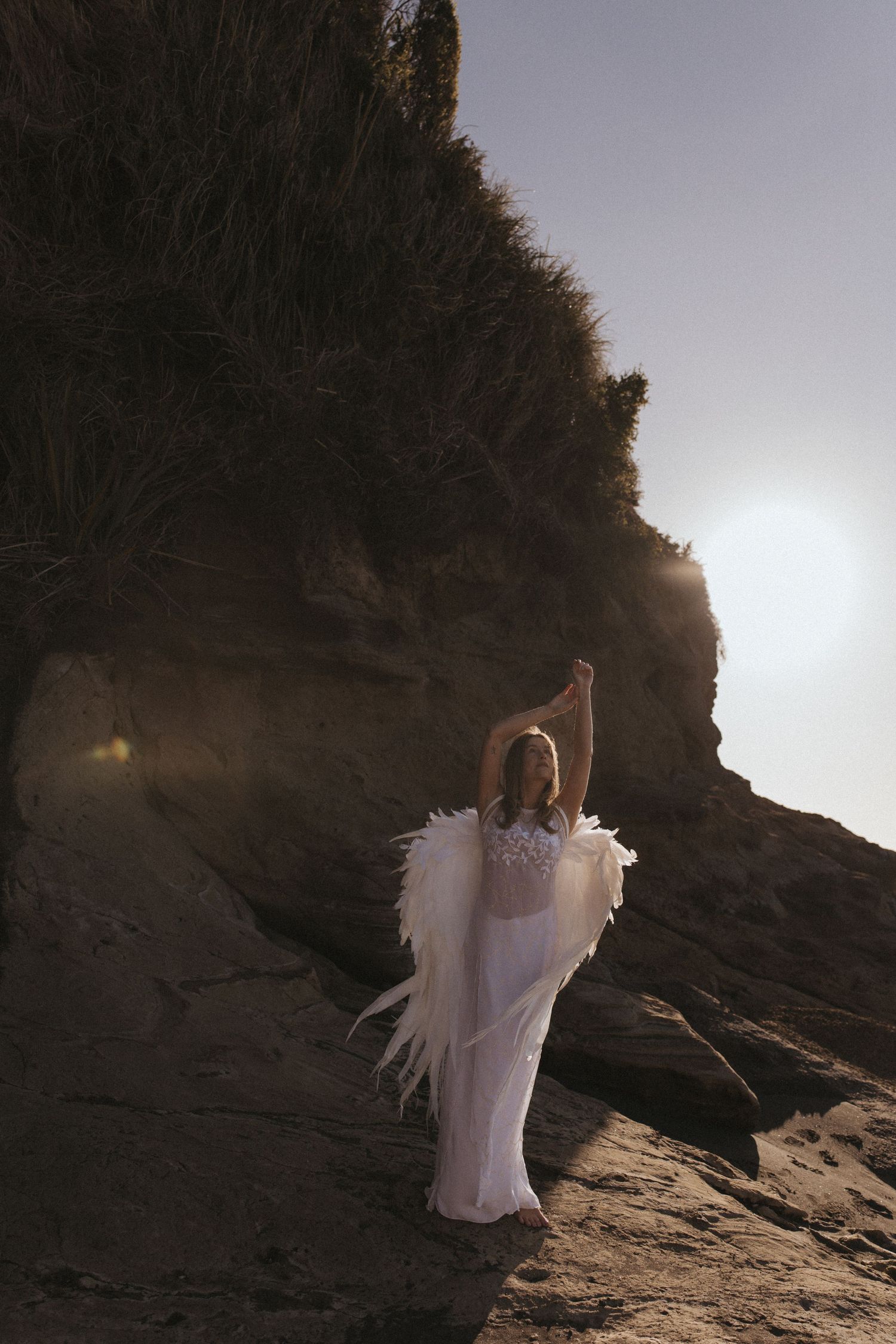 Woman in a flowing white dress and feathered angel wings raises her arms on a rocky coastal cliff at sunset.