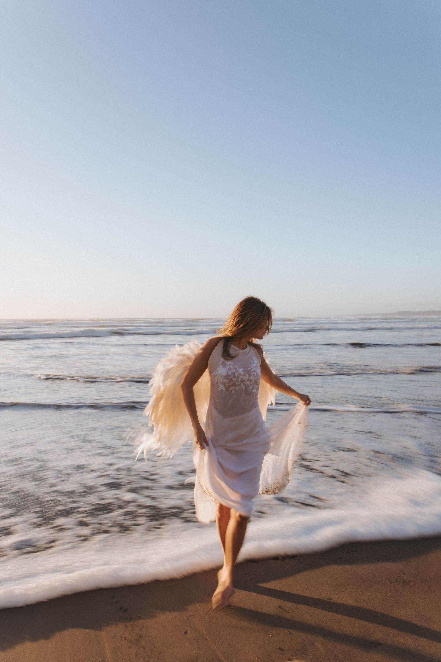 Woman in a flowing white lace dress walks barefoot along the shoreline as ocean waves wash over the sand at sunset.