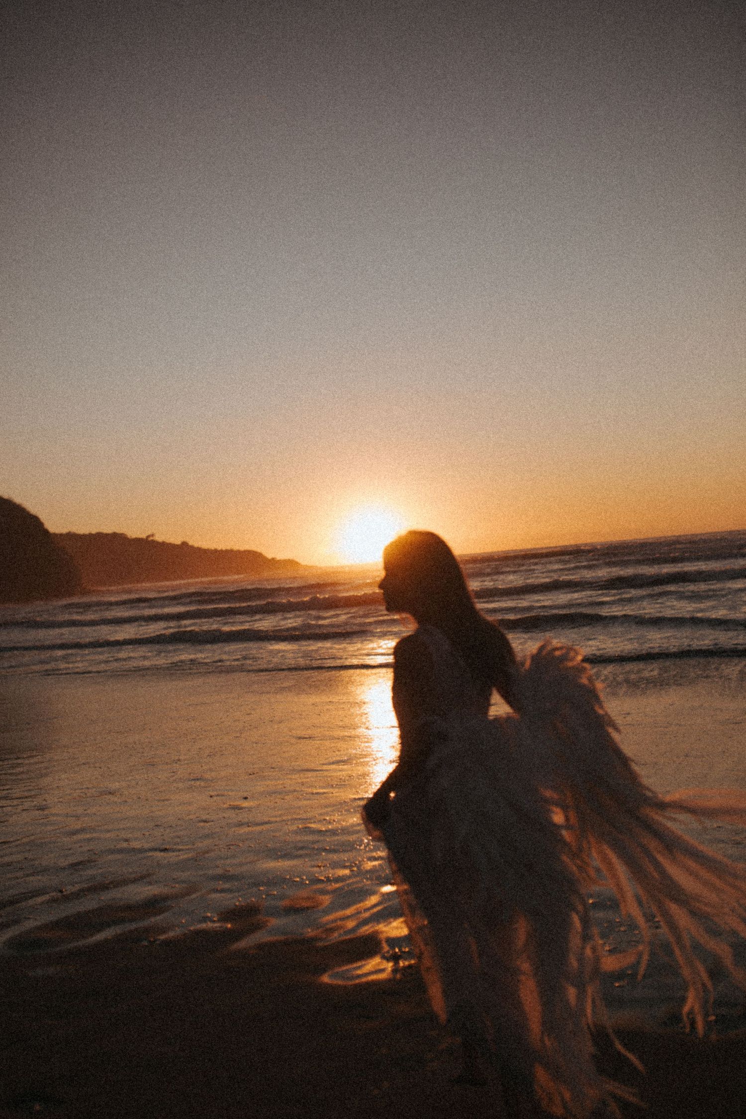 Silhouette of a woman in a flowing feathered dress standing on a wet beach at golden sunset with waves and coastal cliffs behind her.