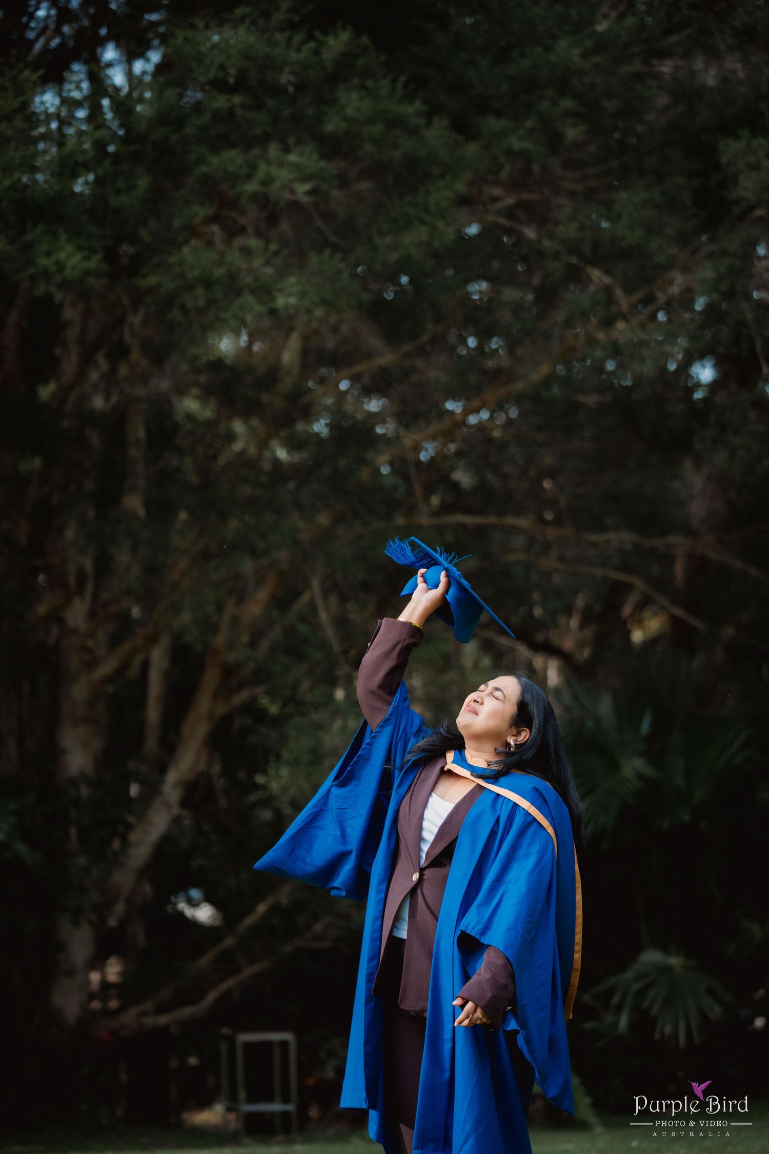Graduate in a blue academic gown tosses her graduation cap into the air while smiling outdoors beneath tall evergreen trees.