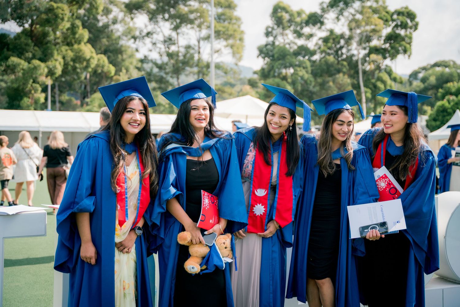 Five smiling female graduates in blue gowns and caps pose together outdoors at a sunny university graduation ceremony.