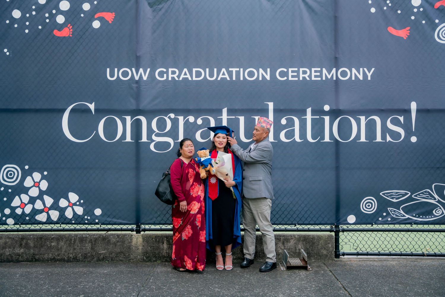 A graduate in a blue gown and cap poses with two family members in front of a UOW Graduation Ceremony Congratulations banner.