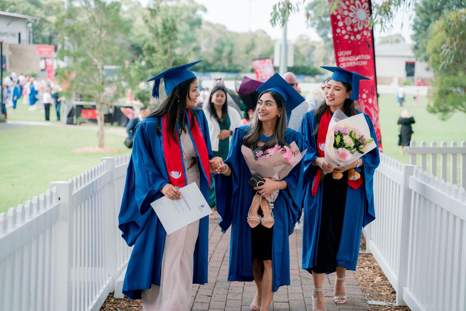 Smiling graduates in blue gowns and caps walk together along a path holding bouquets of flowers and diplomas on a sunny campus.