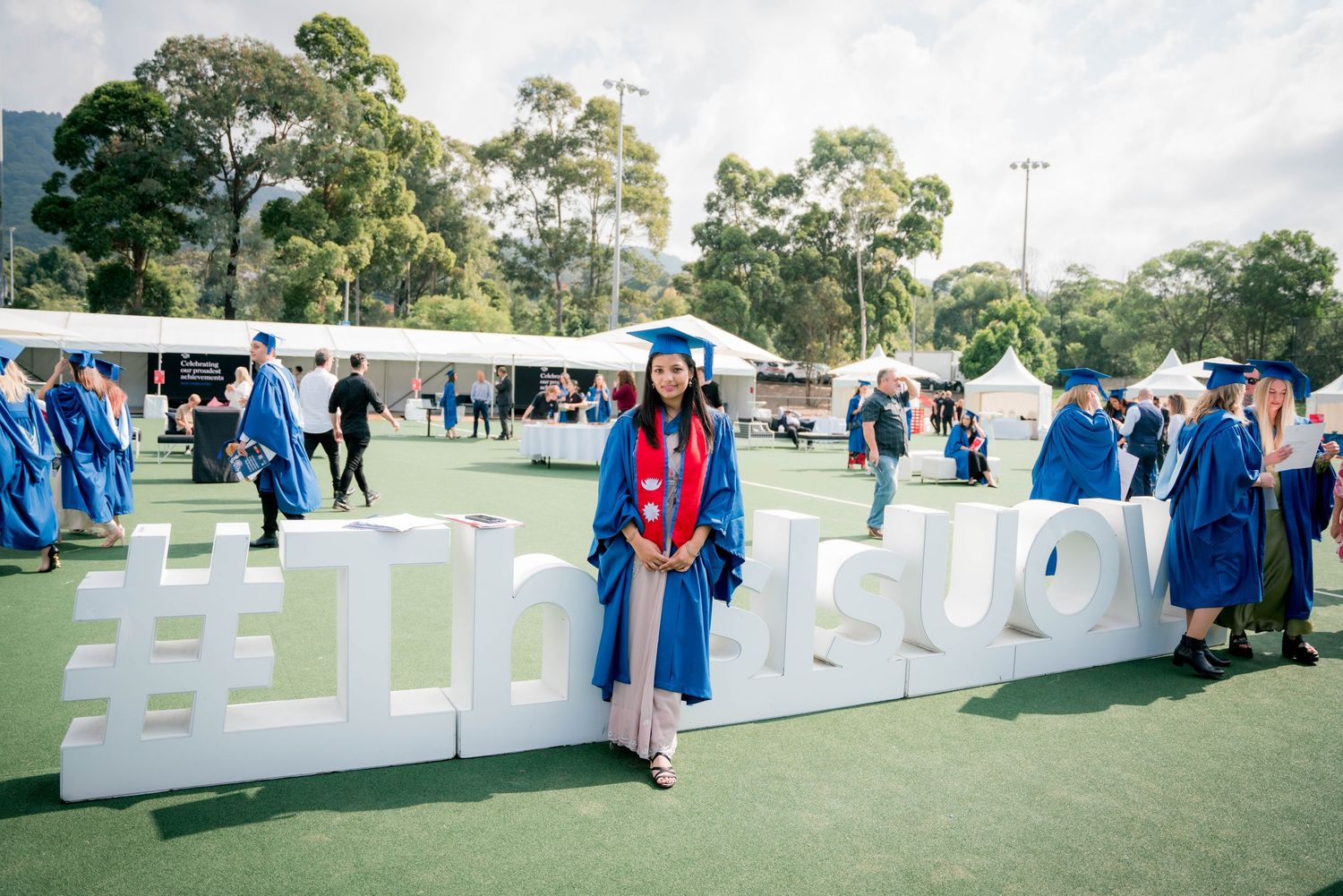Graduate in blue cap and gown with red stole stands beside large white hashtag letters on a grassy lawn at an outdoor commencement ceremony.