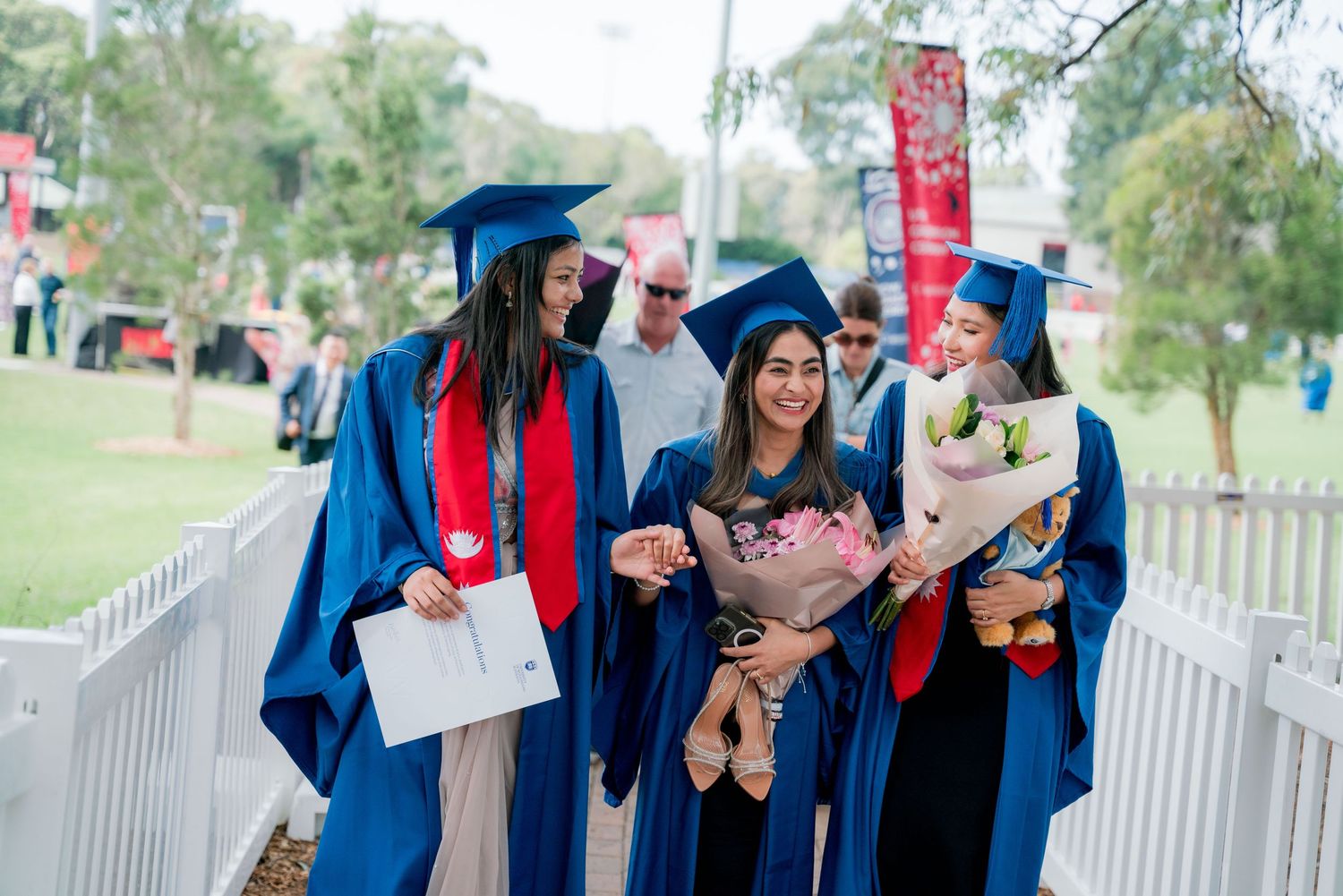 Three smiling graduates in blue caps and gowns walk outdoors holding bouquets of flowers and a diploma folder on graduation day.