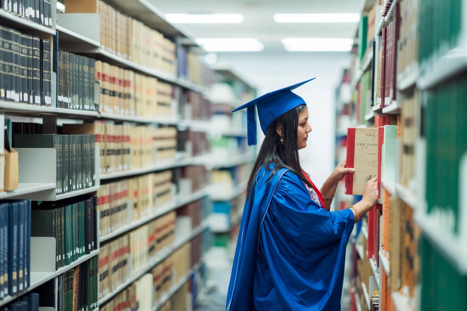 Graduate in a blue cap and gown browses books on tall library shelves filled with academic volumes.