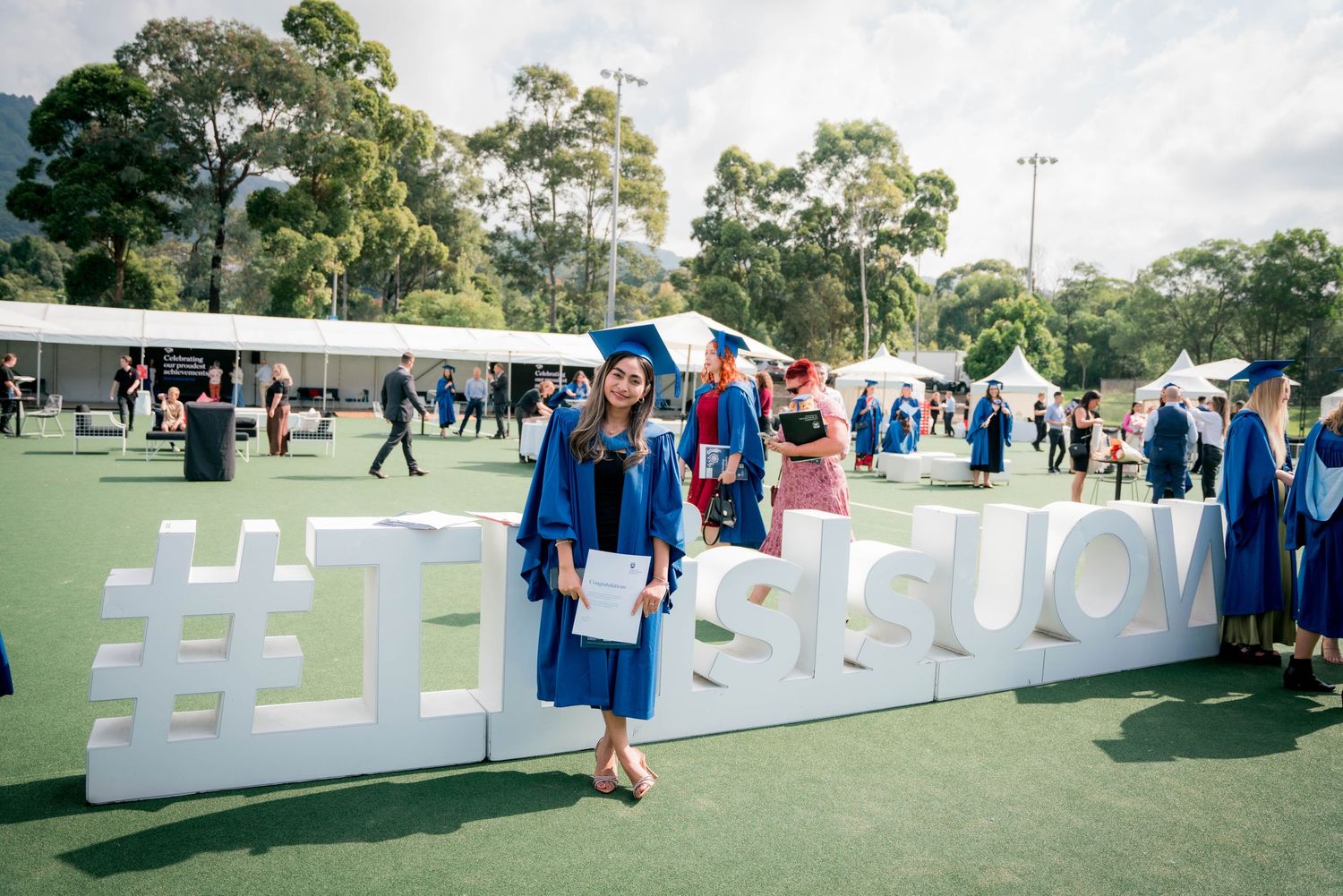 Graduate in a blue gown and cap holding a diploma beside large white hashtag letters on a sunny lawn, with graduates and tents behind.