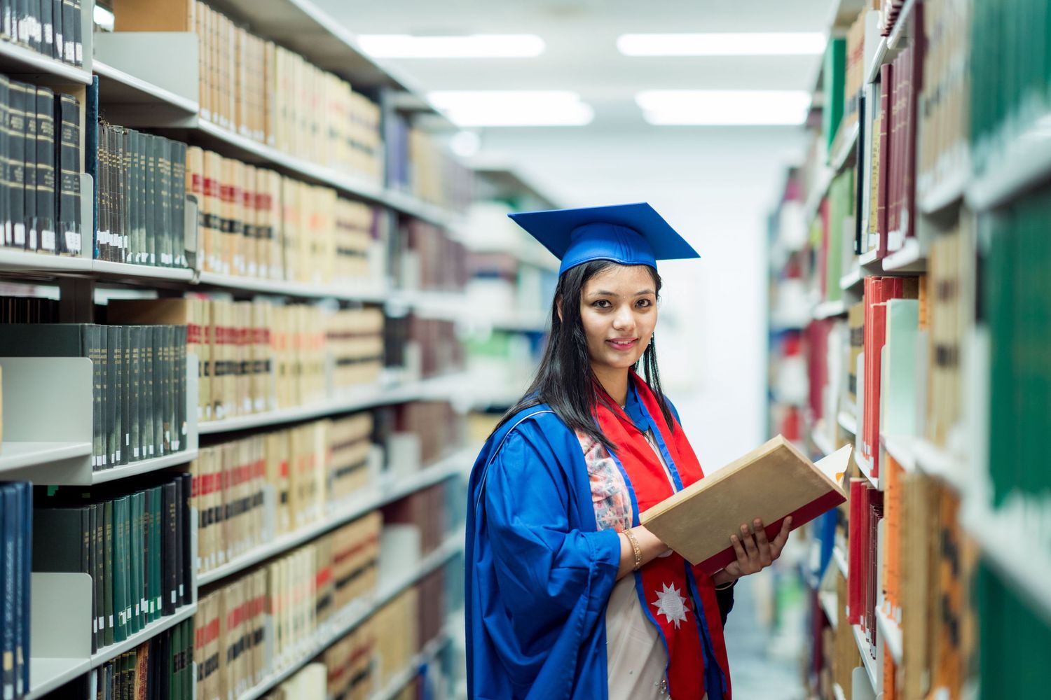 Female graduate in blue gown and cap holds an open book while standing between library shelves at the University of Wollongong.