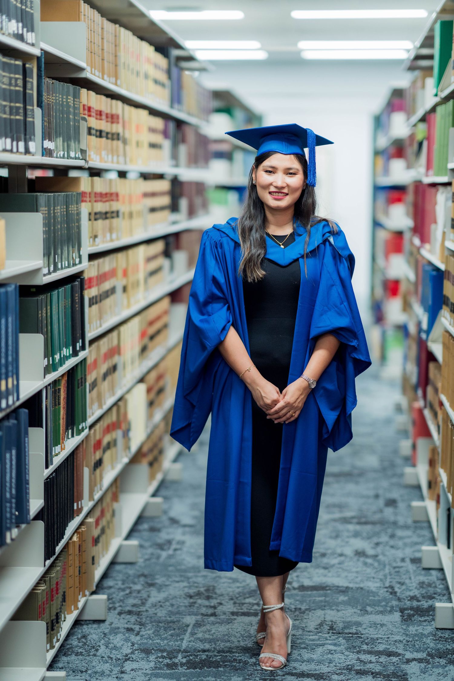 Smiling graduate in a blue gown and cap stands with clasped hands between tall library bookshelves.