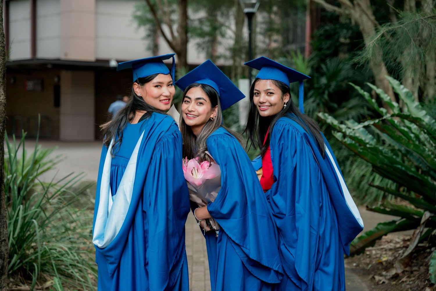 Three smiling graduates in blue gowns and caps pose together outdoors, one holding a pink bouquet, surrounded by green tropical plants.