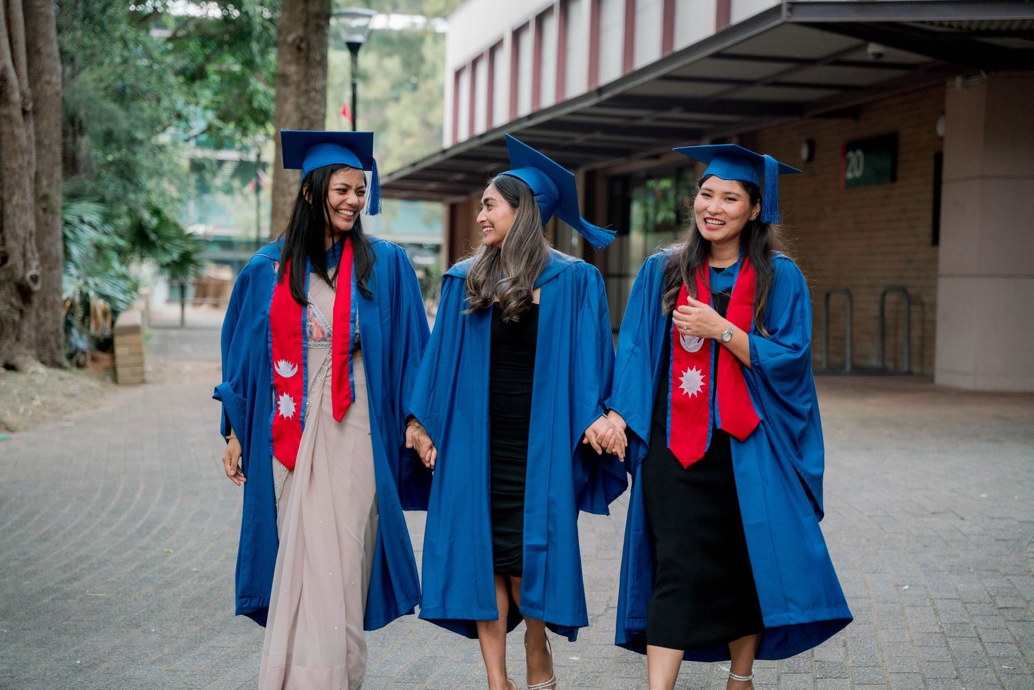 Three smiling female graduates in blue gowns and caps walk hand in hand outside a university building on campus.