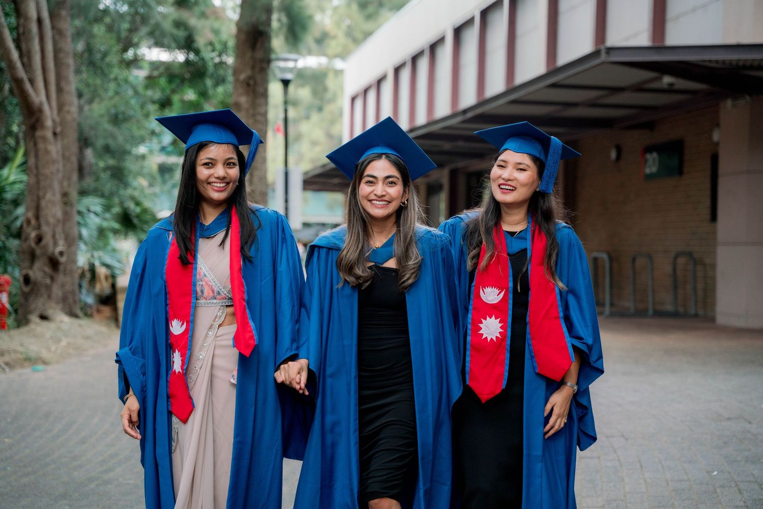 Three smiling graduates in blue gowns, caps and red stoles walking arm in arm outside a campus building.
