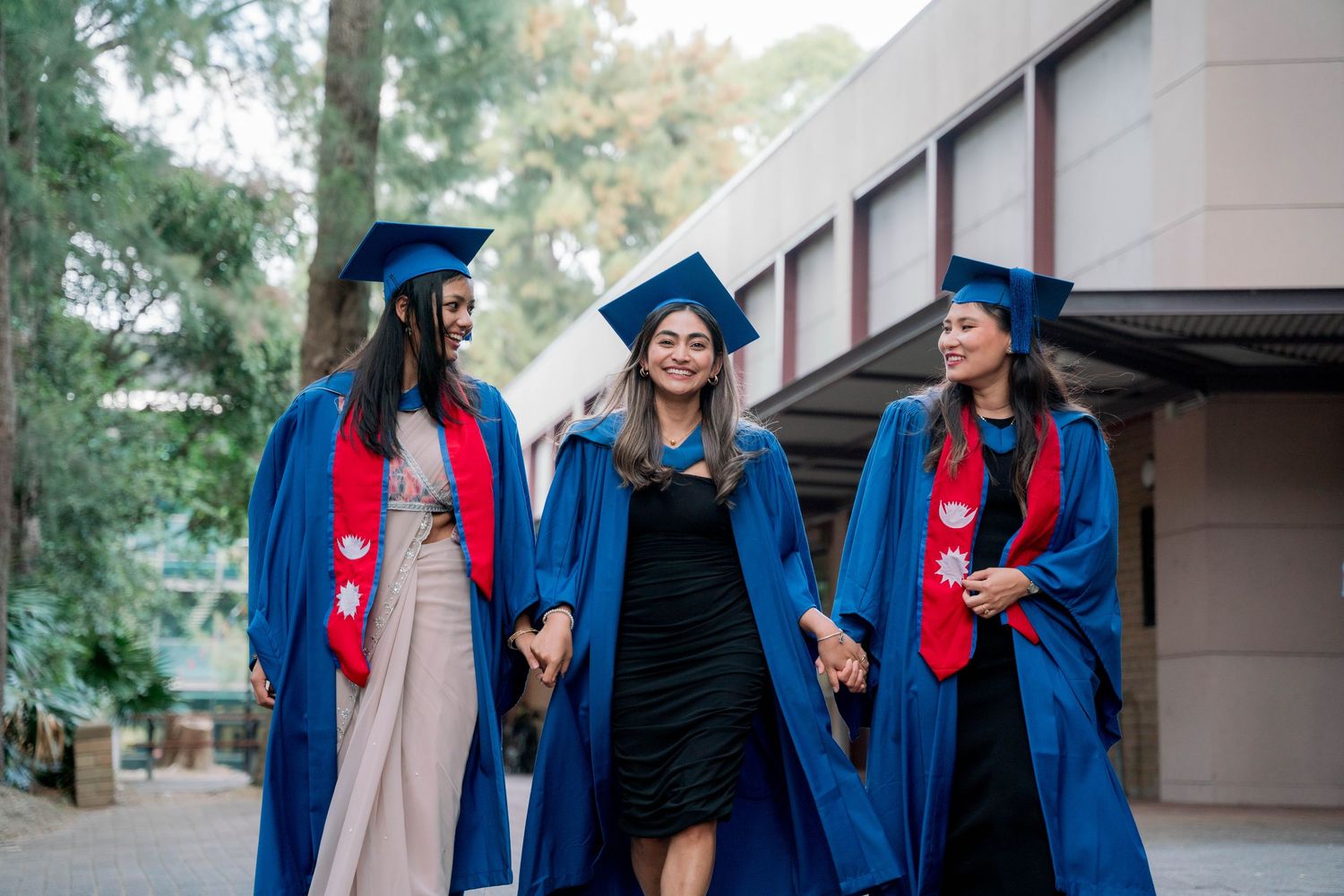 Three female graduates in blue gowns and red stoles walk together holding hands past a campus building surrounded by trees.