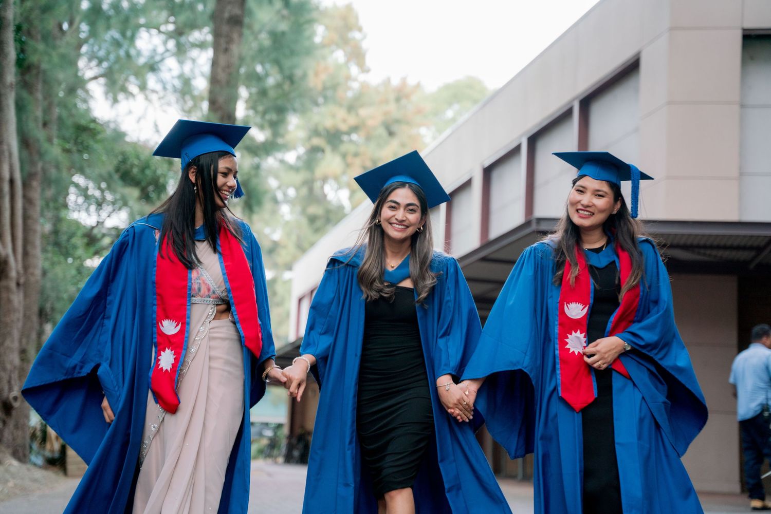 Three smiling female graduates in blue gowns and caps hold hands while walking outdoors near a university building on graduation day.