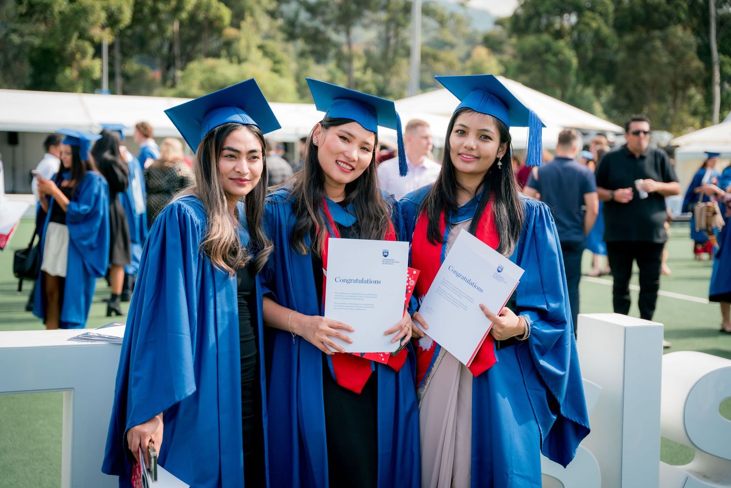 Three smiling graduates in blue gowns and caps hold their diplomas at an outdoor graduation ceremony with guests and tents behind them.