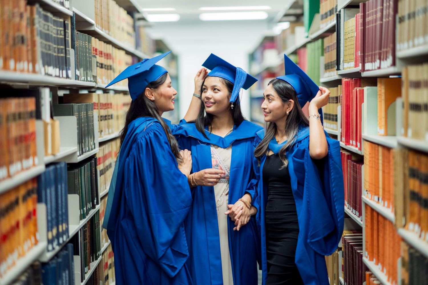Three female graduates in blue gowns and caps smile and embrace between tall library bookshelves filled with books.