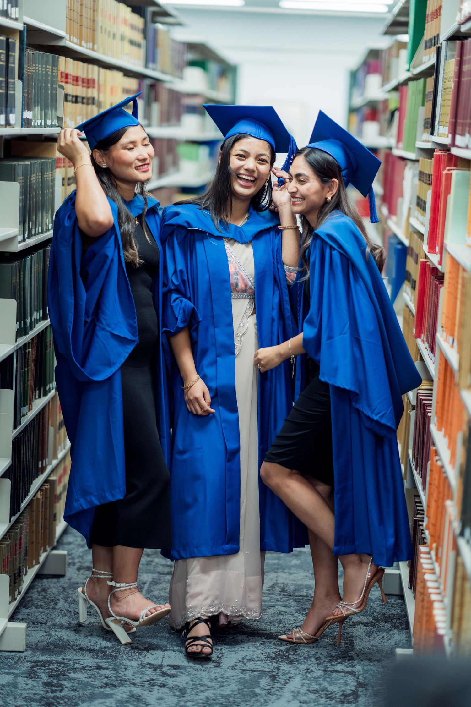 Three joyful female graduates in blue gowns laugh and pose together between tall library bookshelves.