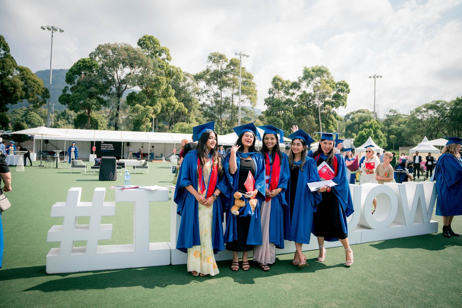 Group of graduates in blue gowns and caps smiling for a photo beside large white letters on a sunny campus lawn.