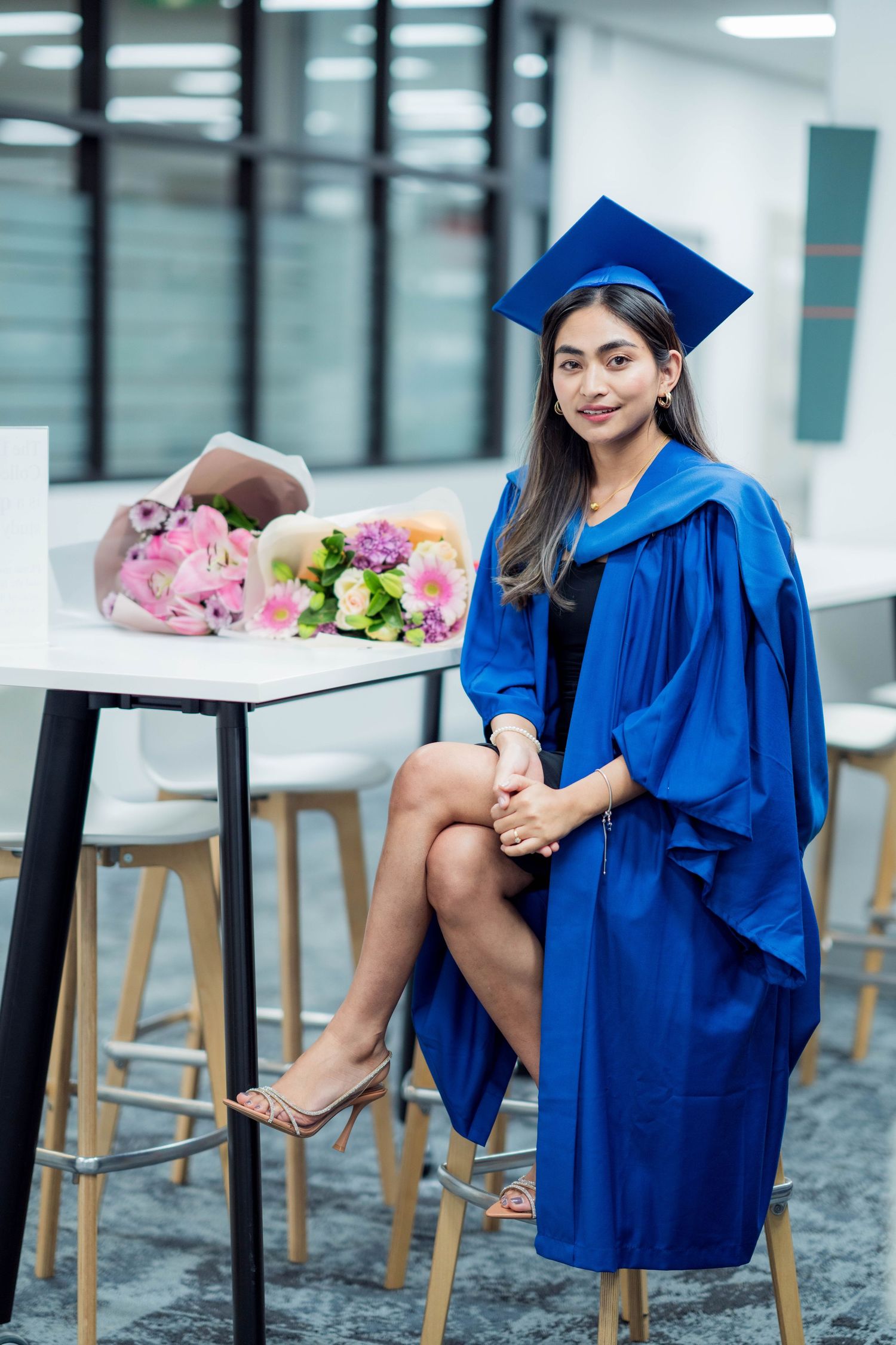 Graduate in a blue cap and gown sits on a stool beside a white table holding two colorful bouquets of pink and purple flowers.