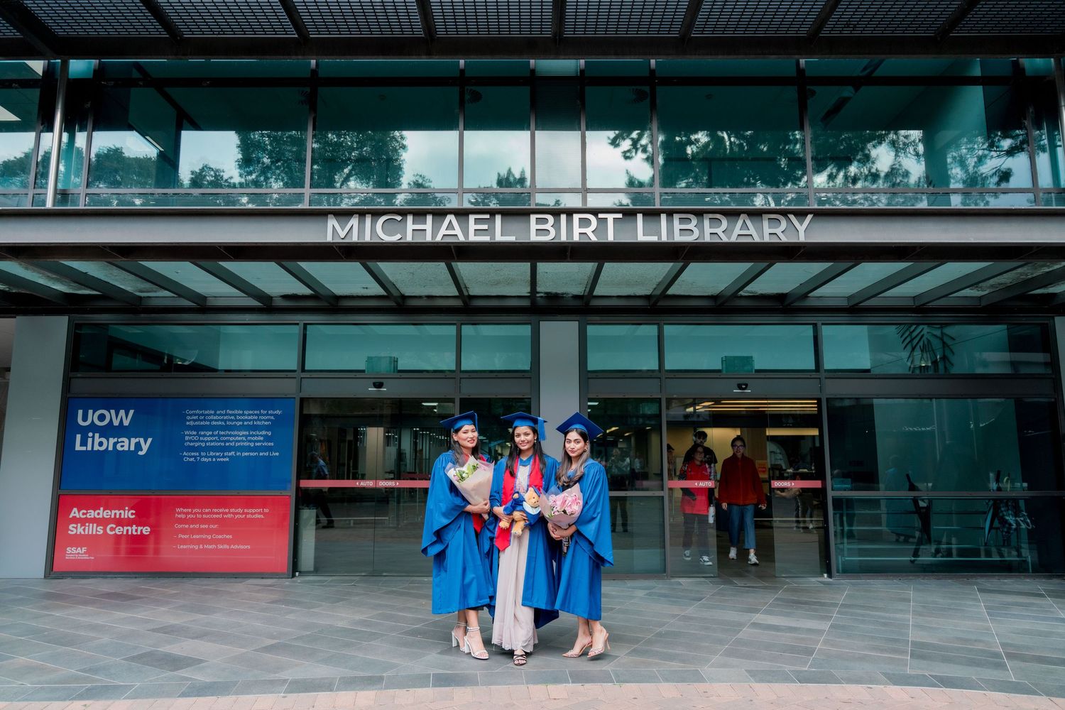 Three graduates in blue gowns and caps hold flowers outside the Michael Bird Library at the University of Wollongong.