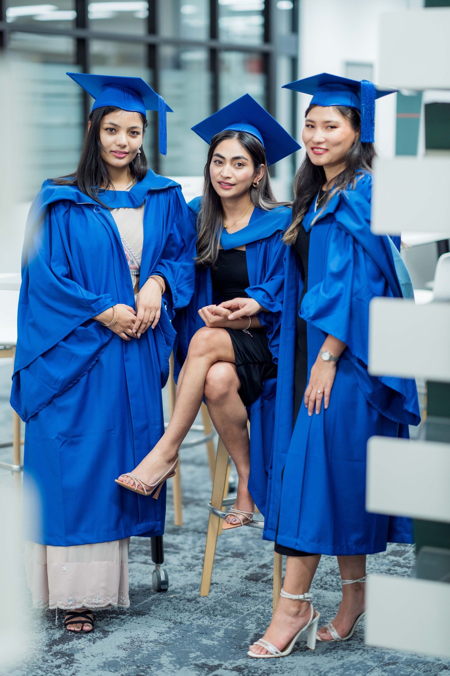 Three smiling women in royal blue graduation gowns and caps pose together indoors, two standing and one seated on a stool between them.