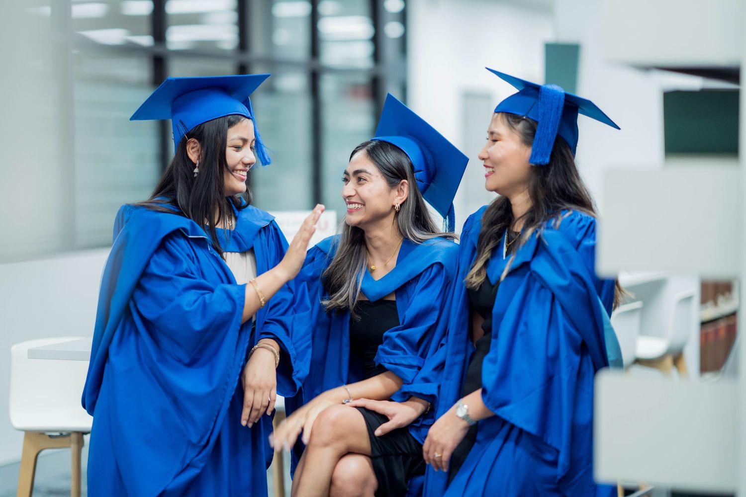 Three female graduates in blue gowns and caps sit together chatting and smiling inside a bright university building.