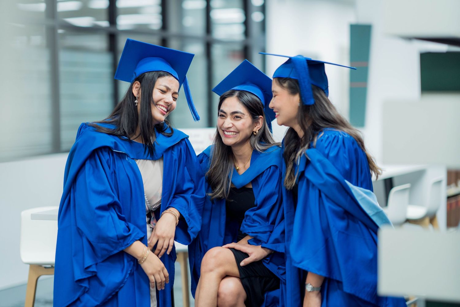 Three female graduates in blue gowns and caps laugh together while seated in a modern university space.