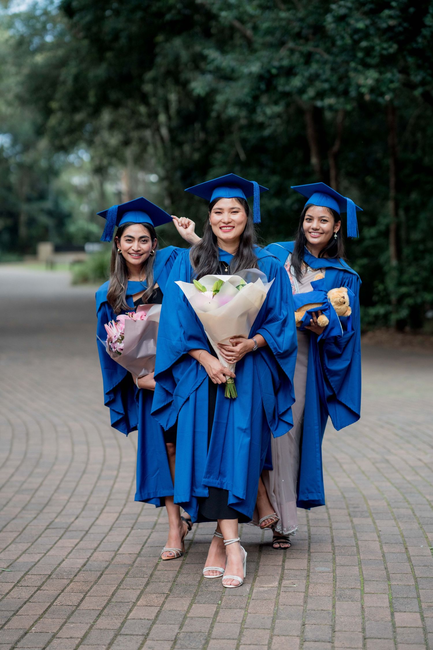 Three smiling graduates in blue gowns and caps hold bouquets of flowers while posing together on a brick path lined with trees.