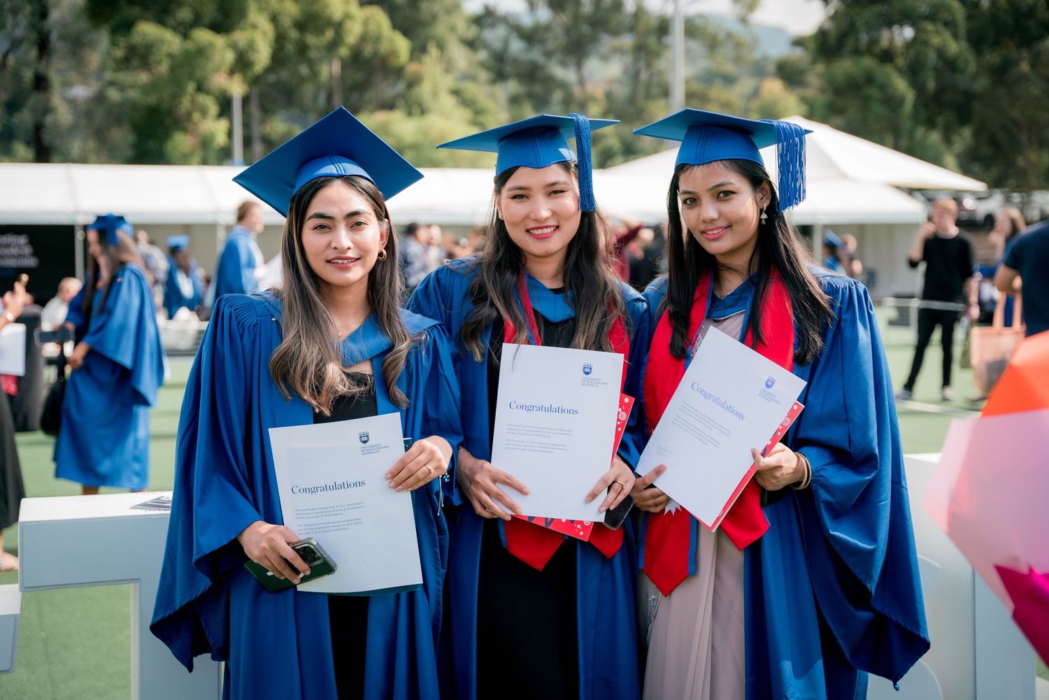 Three smiling female graduates in blue gowns and caps hold their diplomas at an outdoor university graduation ceremony.