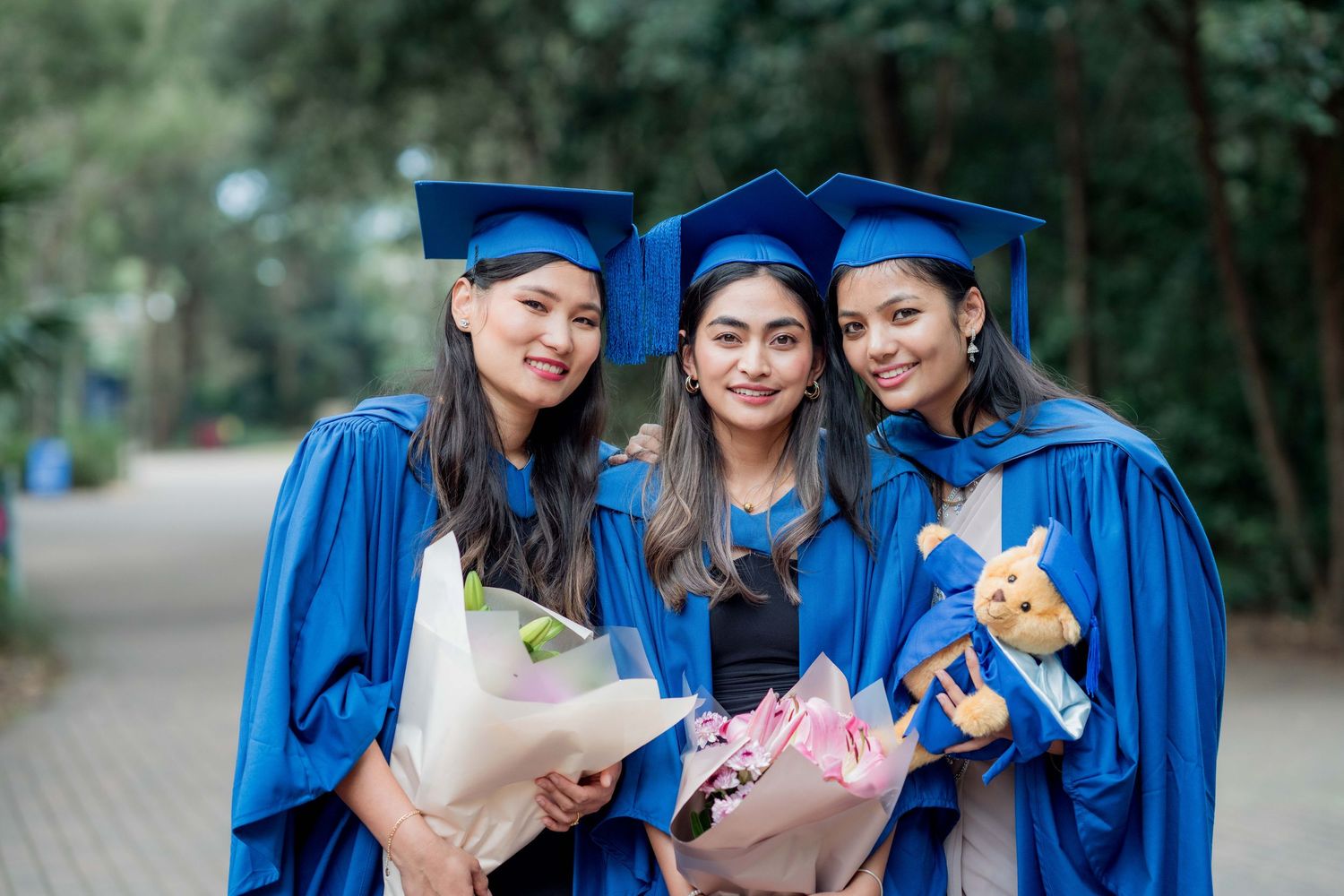 Three smiling female graduates in blue gowns and caps pose outdoors holding flower bouquets and a teddy bear on a tree-lined path.