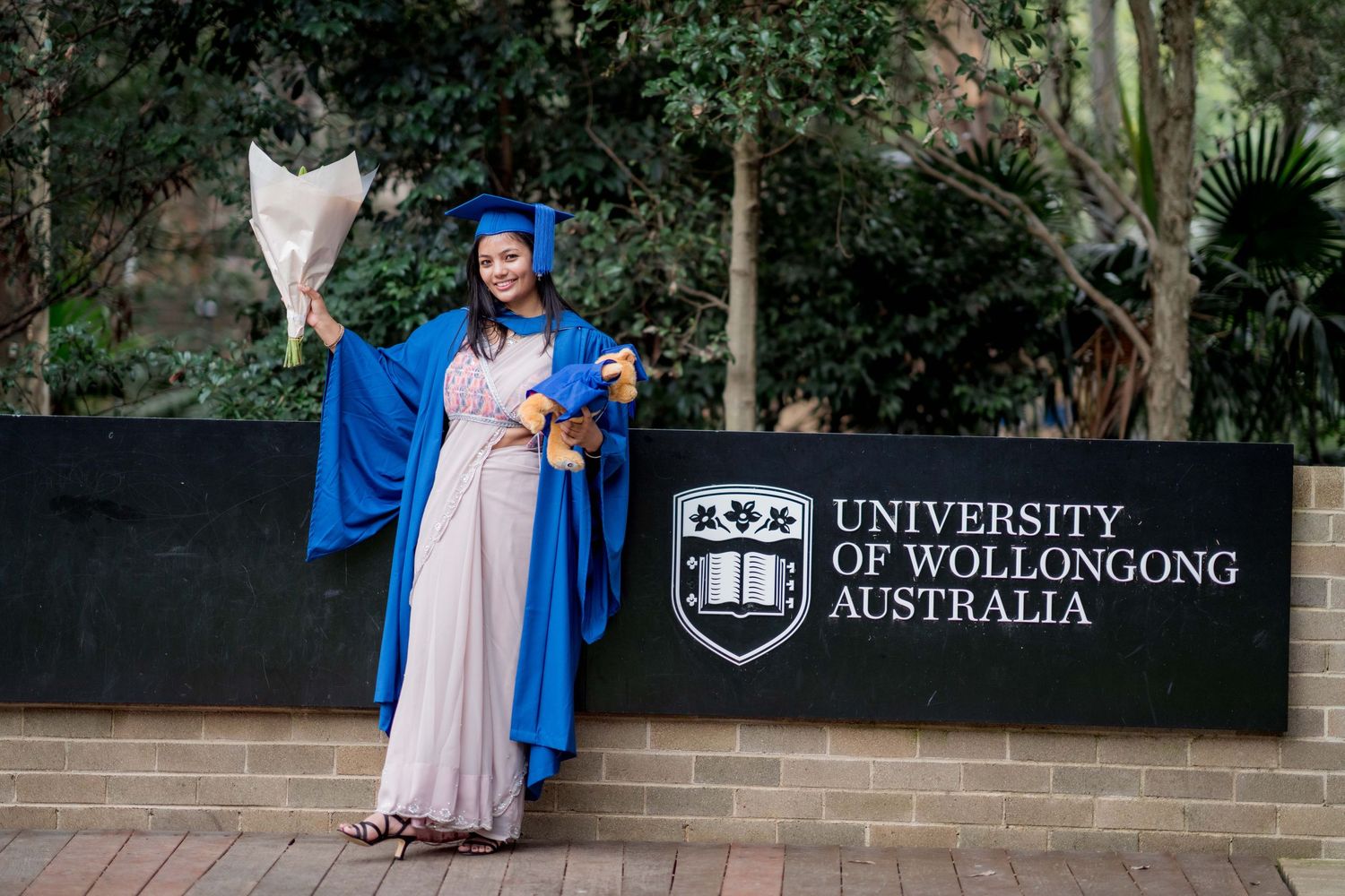 Graduate in a blue gown and cap holds a white bouquet beside the University of Wollongong Australia sign during graduation photos.
