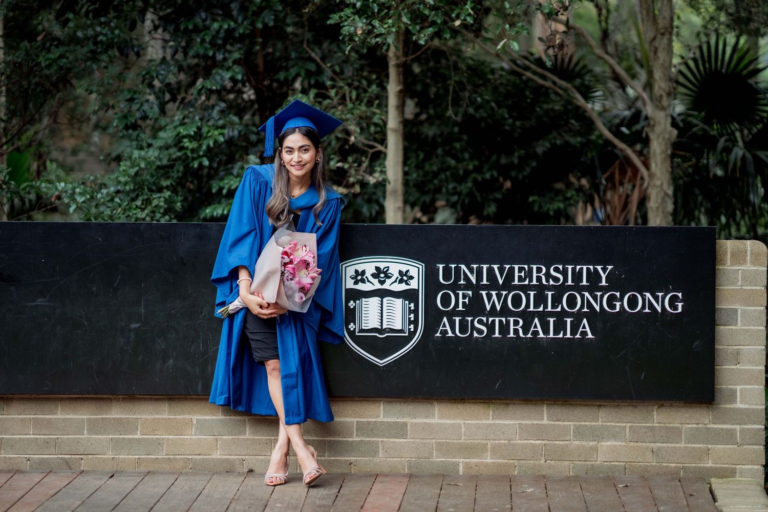 Smiling graduate in a blue gown and mortarboard holds flowers while posing at the University of Wollongong Australia entrance sign.