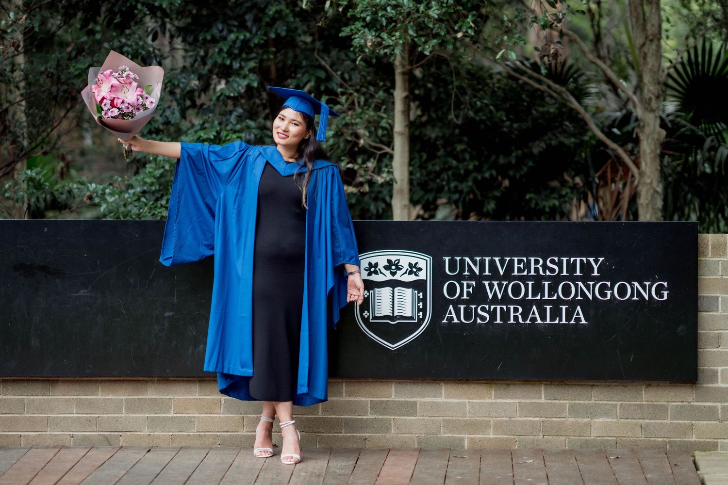 Graduate in a blue gown and cap holding a pink bouquet beside the University of Wollongong Australia sign on graduation day.