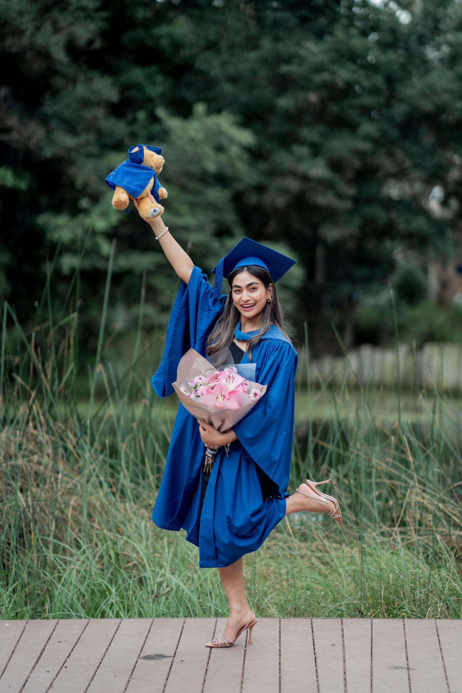 Joyful graduate in a blue gown poses on a wooden boardwalk, holding a teddy bear in a graduation gown and a pink bouquet.