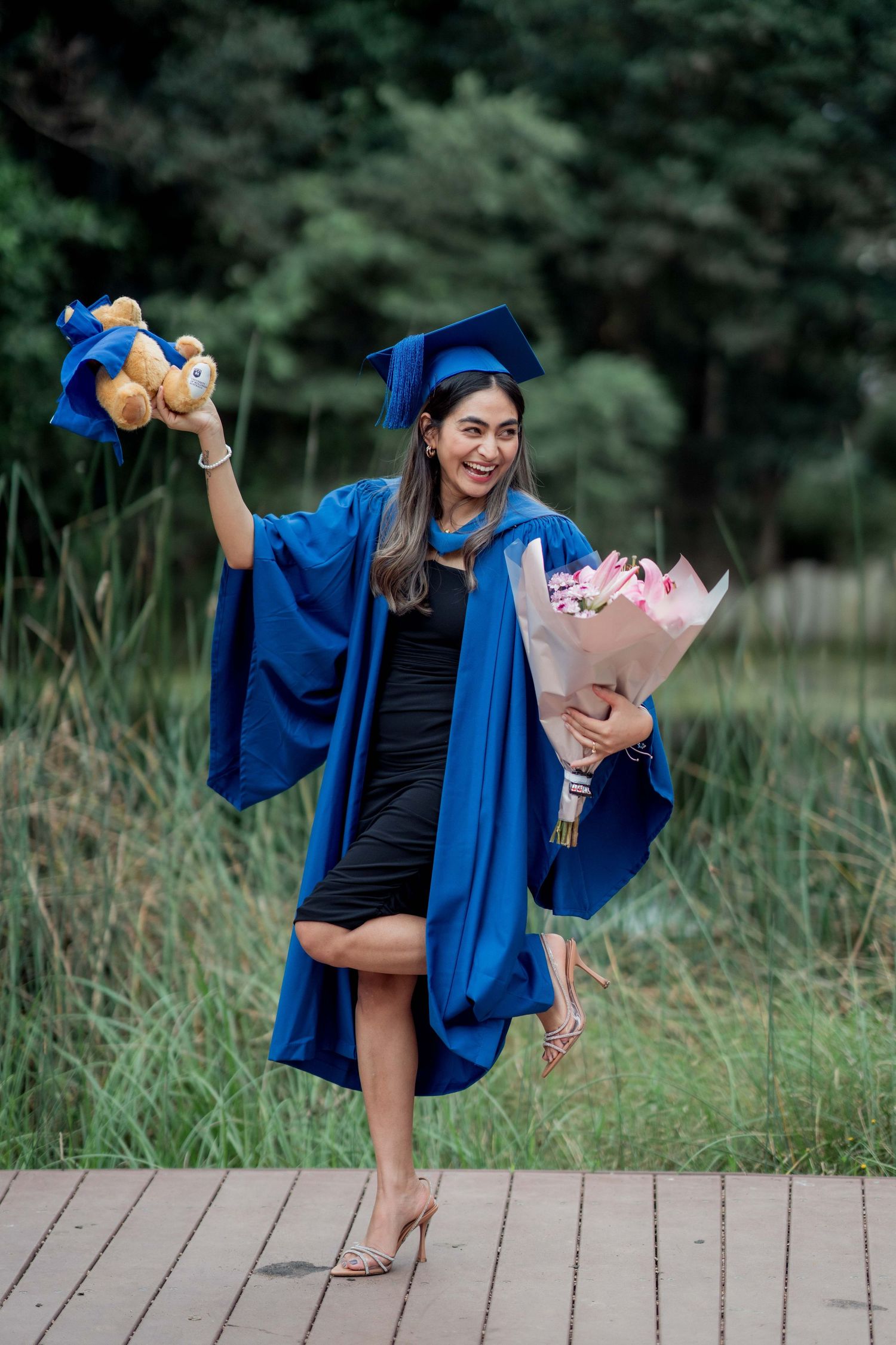 Smiling graduate in a blue gown and cap holds a teddy bear and pink bouquet while posing on a wooden deck near tall green grass.